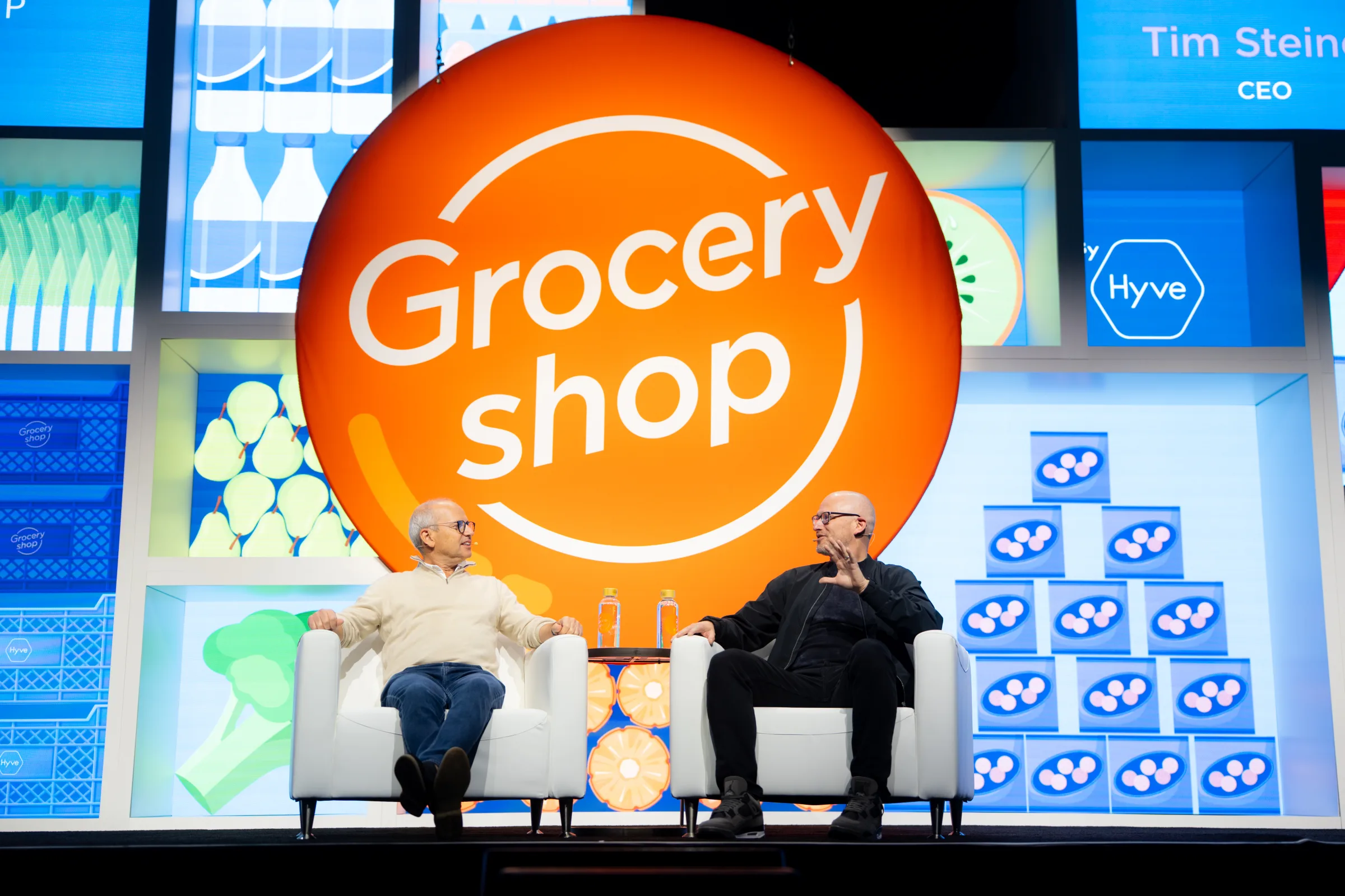 Groceryshop 2025 mainstage: a fireside session framed by the illustrated LED backdrop and the orange logo disc