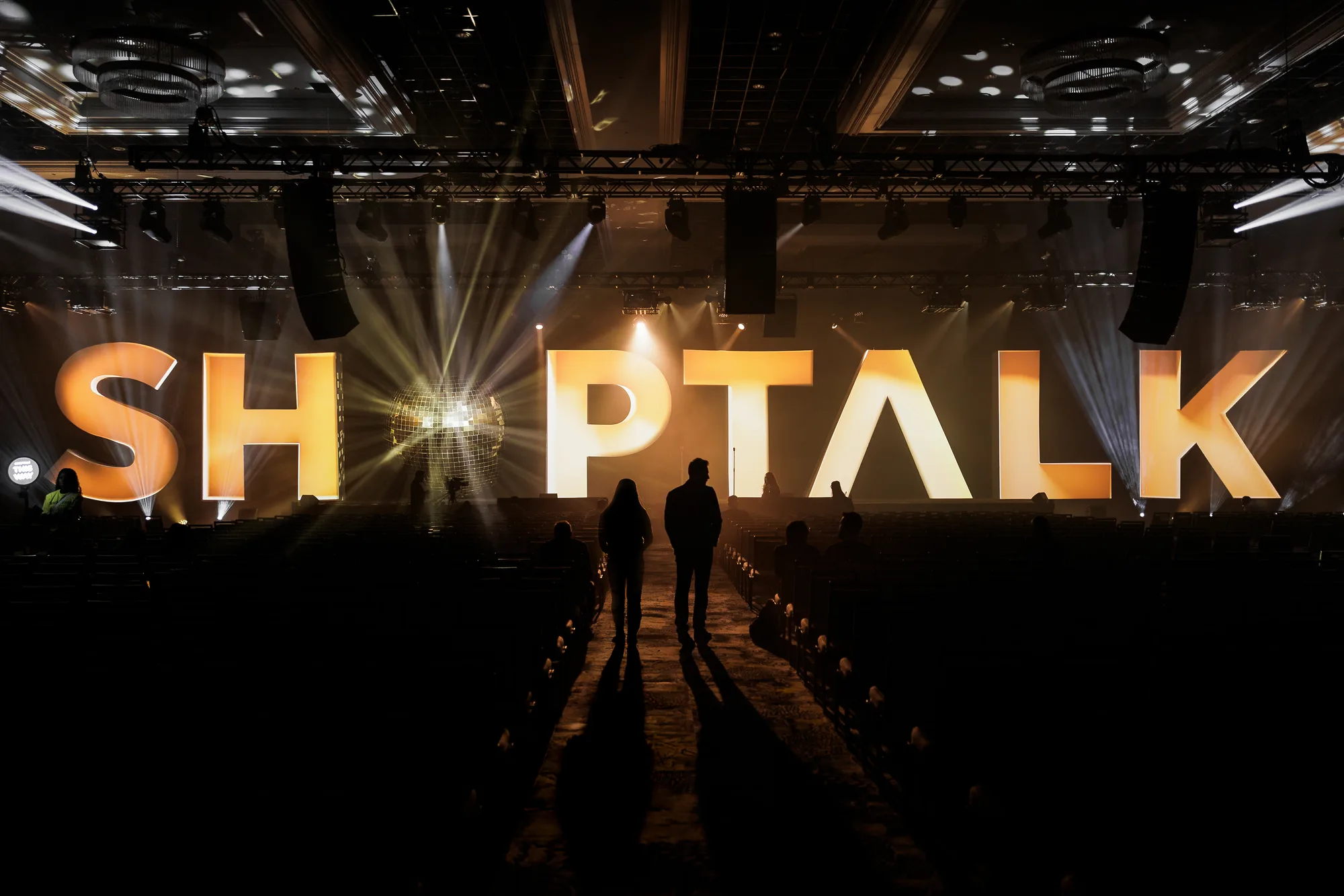 The Shoptalk Spring 2025 mainstage before doors: eight-foot illuminated letters spelling SHOPTALK, a mirror ball set into the O, and two attendees silhouetted in the centre aisle