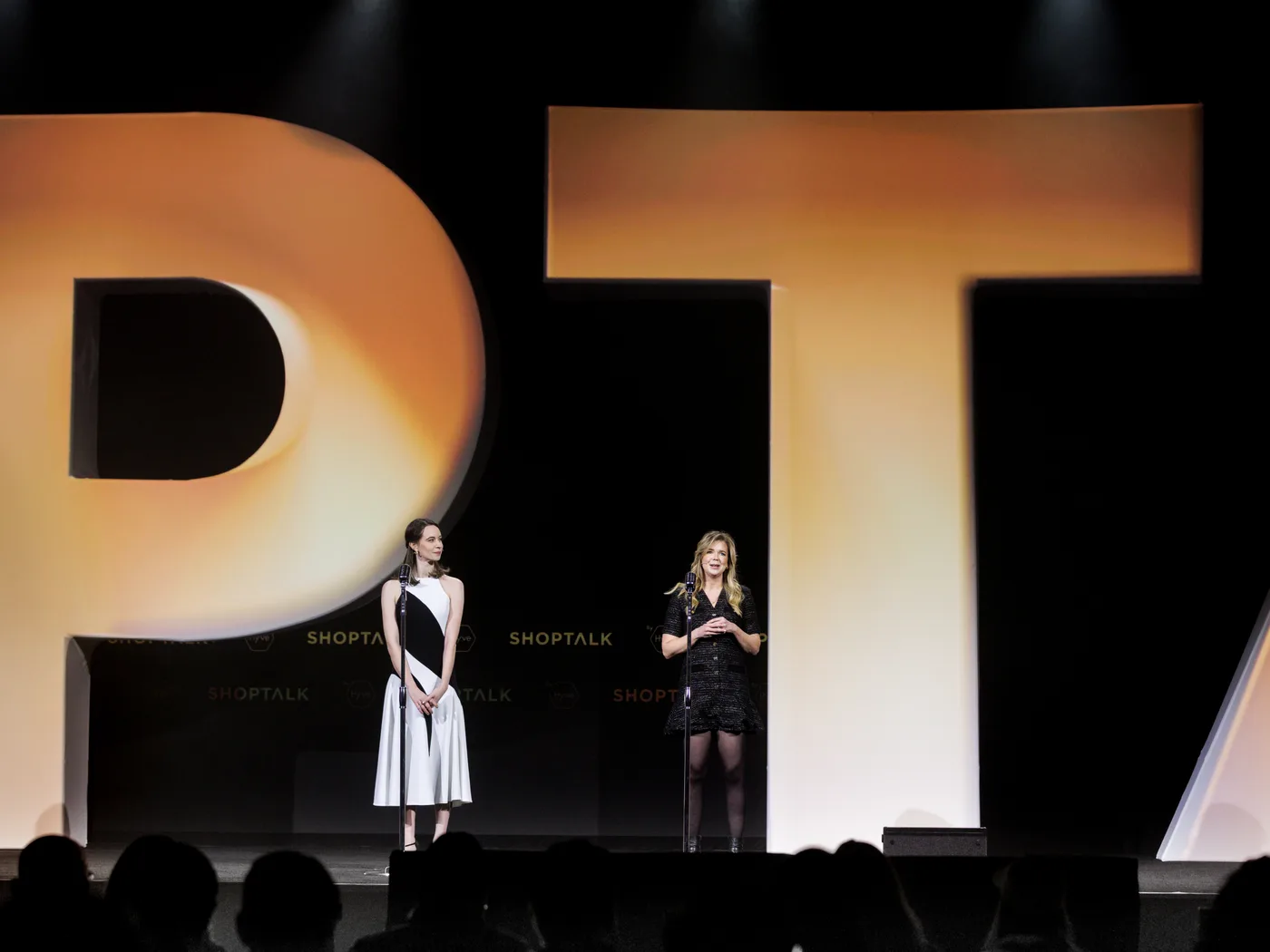 Two speakers standing on the Shoptalk mainstage, dwarfed by the illuminated P and T of the wordmark behind them