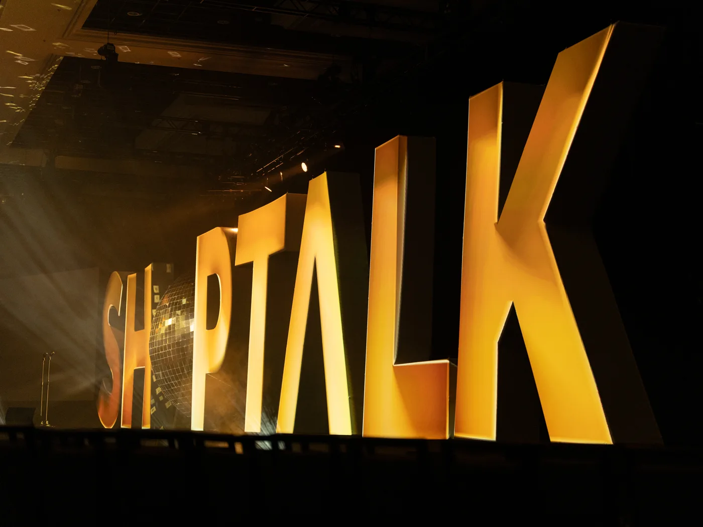 A low raking view along the gold Shoptalk letterforms, showing their built depth, with the mirrored sphere set into the O