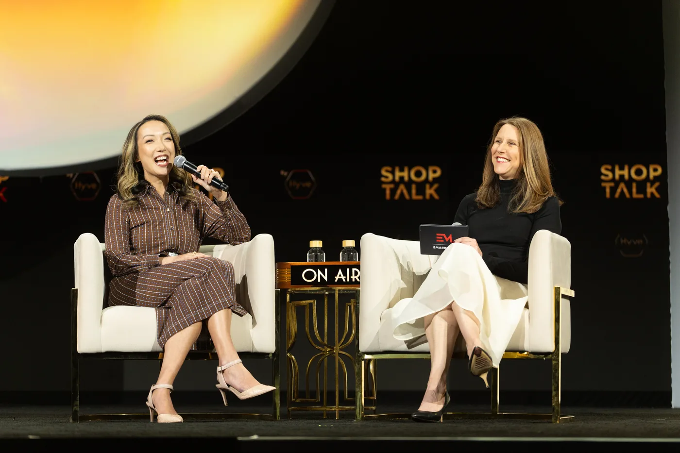 Clara Shih speaking with a microphone on the Shoptalk main stage, the gold sphere glowing above the black back wall