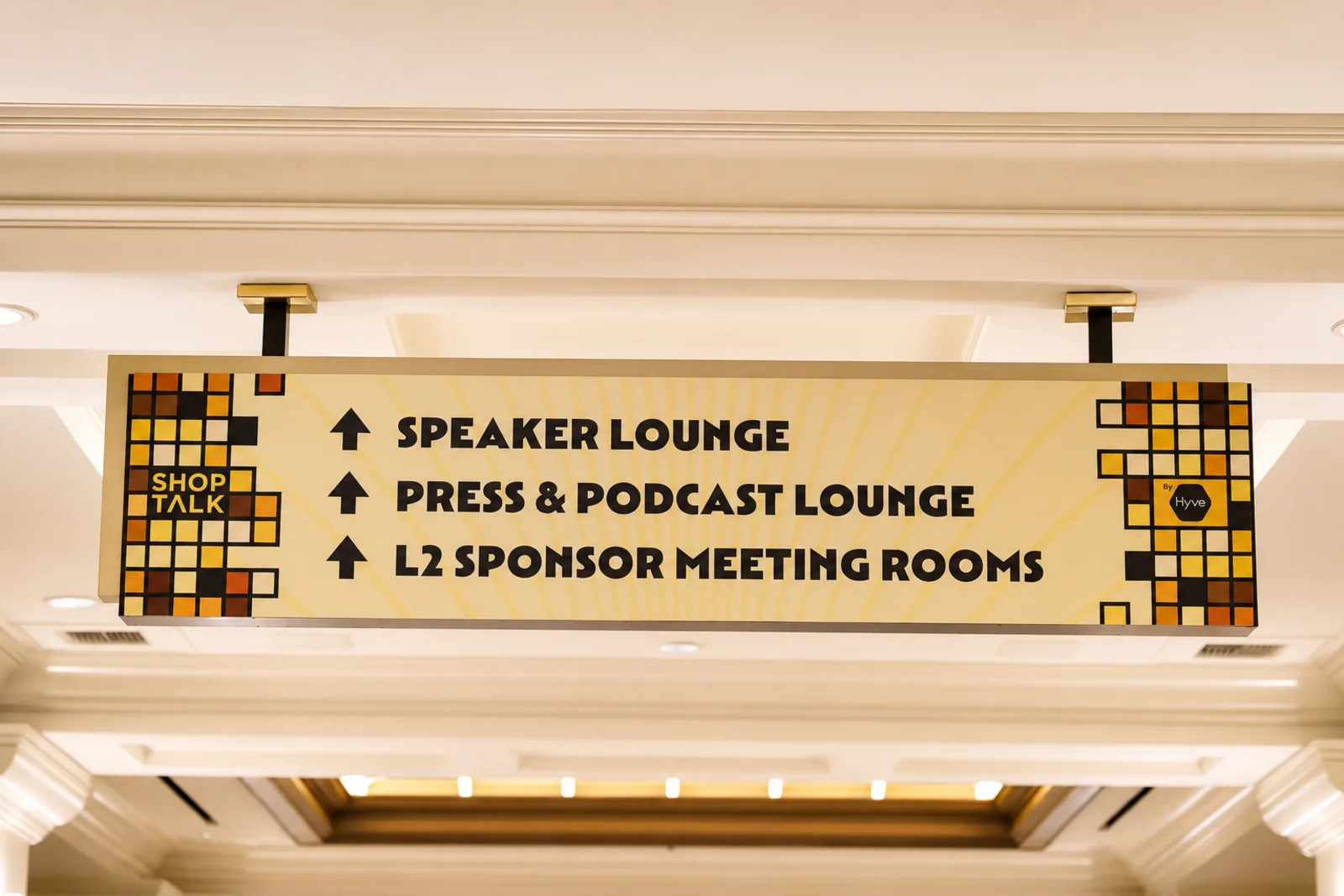 A hanging overhead directional sign in the convention center: mosaic tile blocks at both ends, a pale yellow sunburst ground, and three black arrows pointing up to Speaker Lounge, Press & Podcast Lounge and L2 Sponsor Meeting Rooms