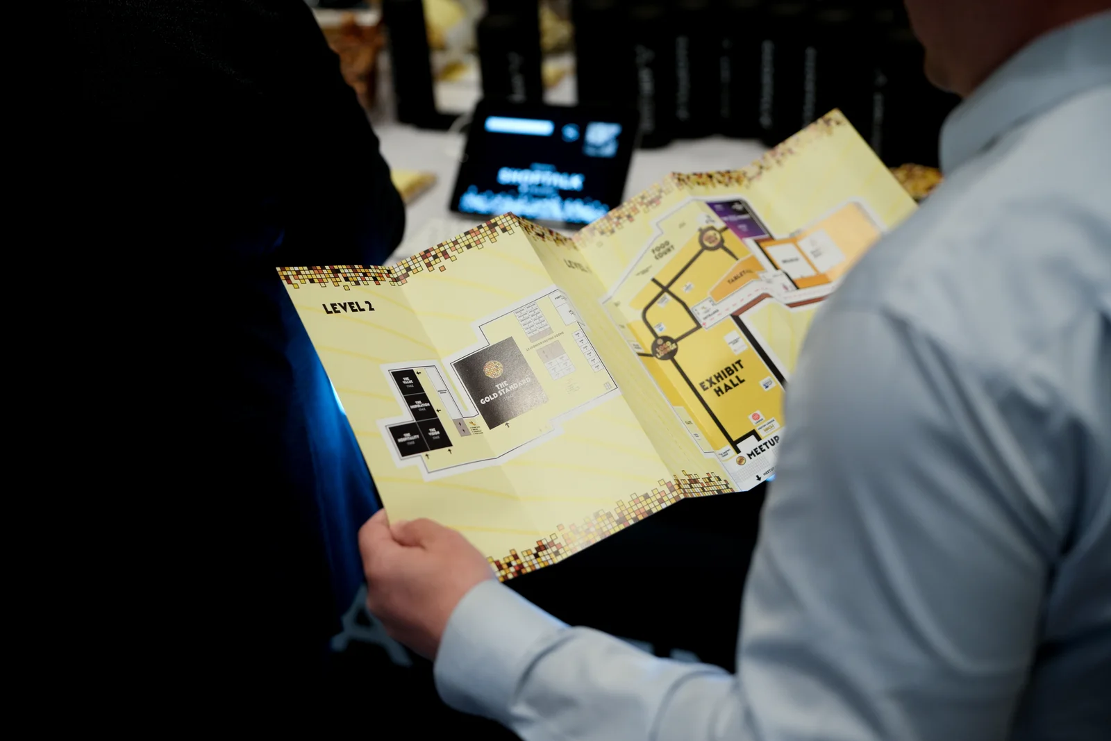 An attendee at the badge desk holding the Shoptalk Spring 2025 printed guide open, the Level 2 stage plan on the left panel and the Level 1 exhibit hall on the right, mosaic tile bands running along the top and bottom edges