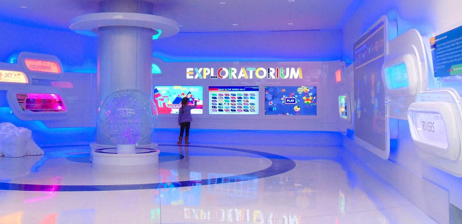The finished Exploratorium waiting room, lit blue, its wordmark glowing above a wall of interactive screens