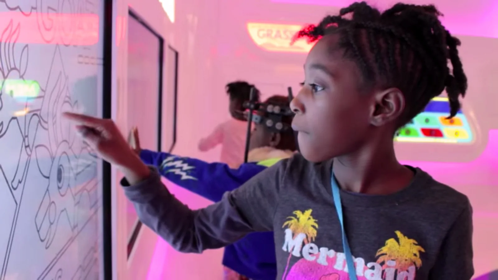 A child tracing a creature outline on a wall touchscreen, the biome panels lit pink behind her