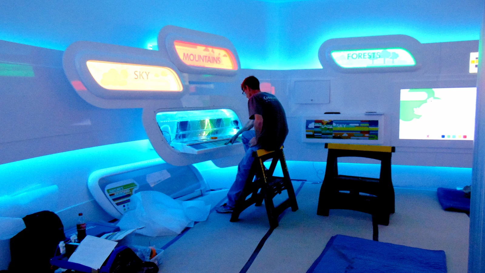 The Exploratorium during installation, backlit biome panels glowing blue and amber around the room